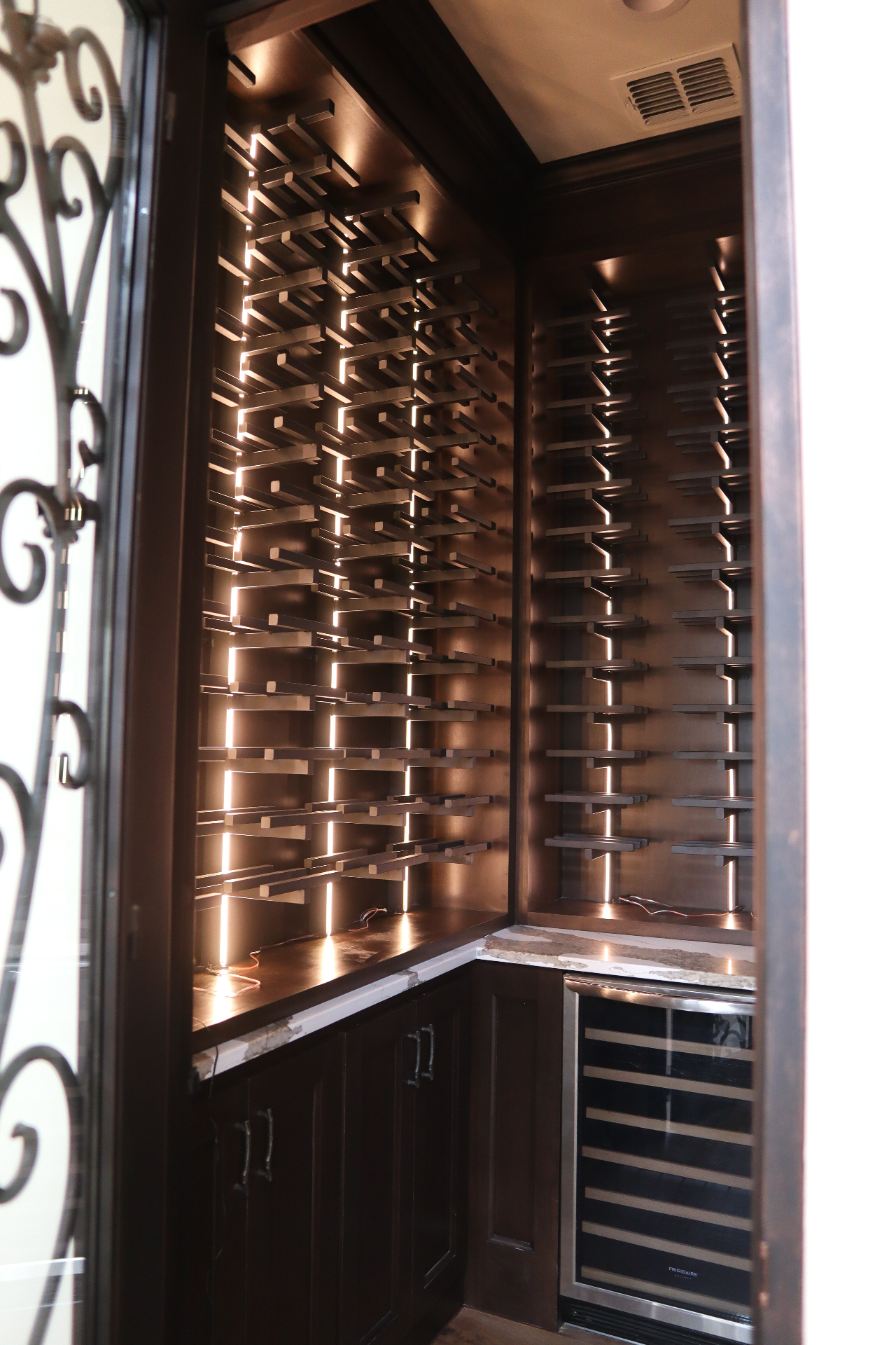 Custom walk-in wine room with stained walnut display racks, integrated lighting, and stone counters by Amaya