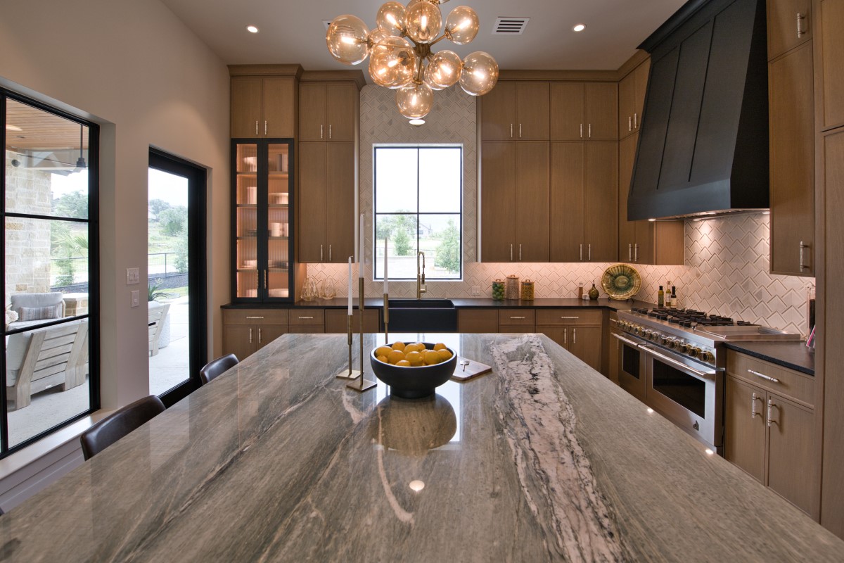 Warm white oak kitchen with obsidian black hood, herringbone backsplash, and a dramatic glass-globe chandelier over a stone island by Amaya